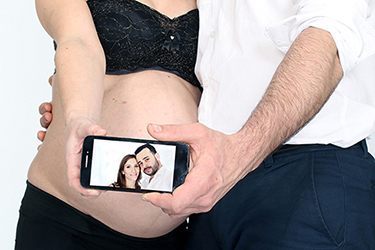 grossesse heureuse photographiée par notre photographe Rachel Joubi, couple montrant leur portrait sur un smart phone qui est placé devant le ventre bombé de la futur maman.