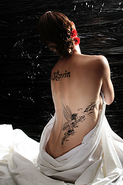 Femme prise de dos nue assise sur un drap avec ses deux tatouages visibles, l'un des tatouages représente une fée et l'autre, il est écrit avec une jolie typo manuscrite Marvin. photographiée par notre photographe Rachel Joubi
